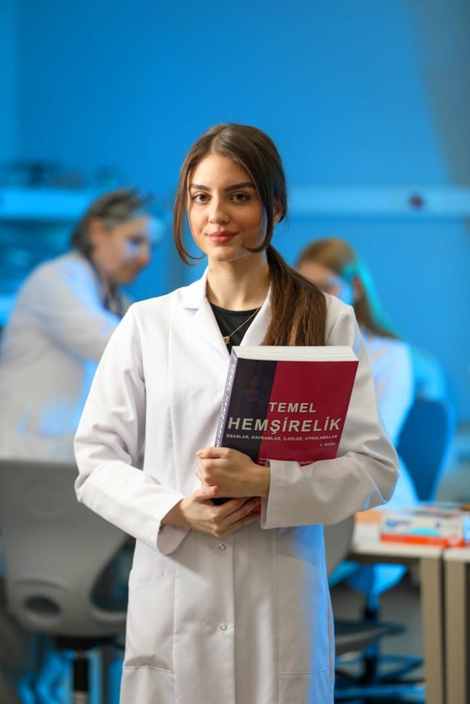 Young woman in a lab coat holding a nursing textbook in a classroom setting.